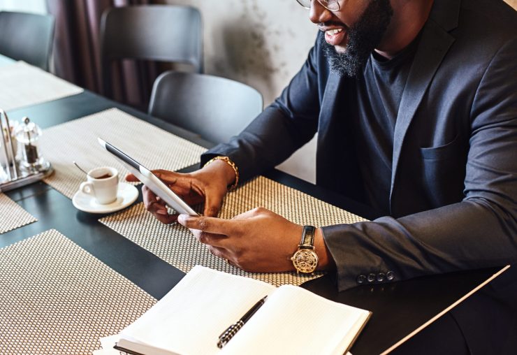 Portrait of focused African-American office manager in dark-blue jacket and glasses, sitting at cafe table with tablet pc, reading documents and smiling. Coffee is on the table. Cozy interior